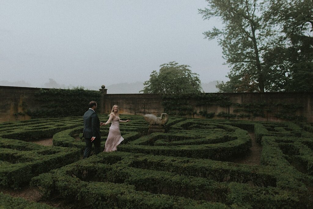 Woman in blush dress turns back and holds onto her fiance's hand as they run through Allerton Park's hedge maze. 