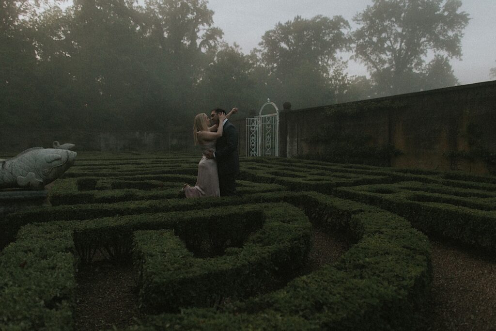 Couple holds each other in Allerton Park's hedge maze surrounded by foggy mist in romantic engagement portraits. 