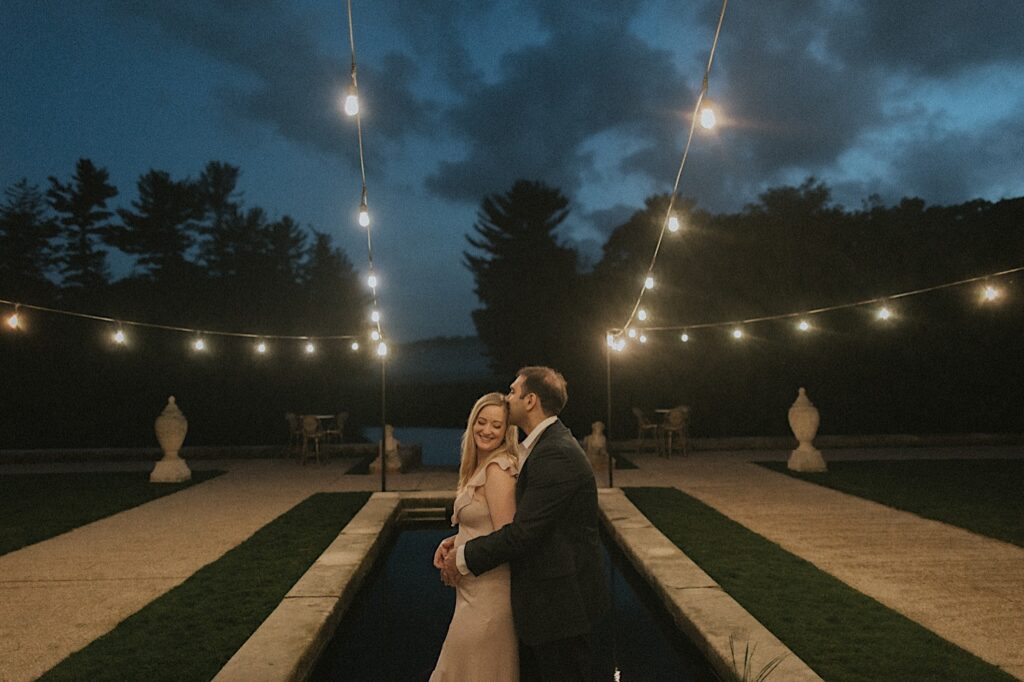 Woman stands while being hugged from behind by her fiance as they stand on the patio of Allerton Park's beautiful mansion lit by patio lights. 