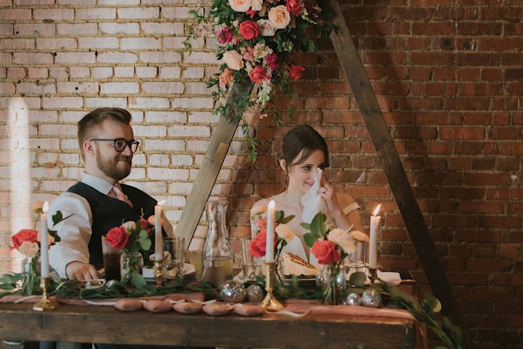 Bride wipes away a tear and sits next to the groom at the head table during wedding reception at Reality on Monroe in Central Illinois. 