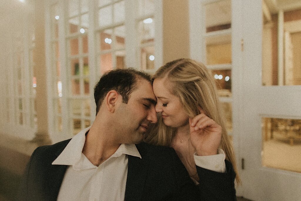 Couple places their heads together as they sit in front of a window in a romantic engagement portrait. 