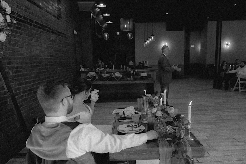 Bride and groom sit at the head table as they look at the father of the bride giving a speech to the guests at the reception. 