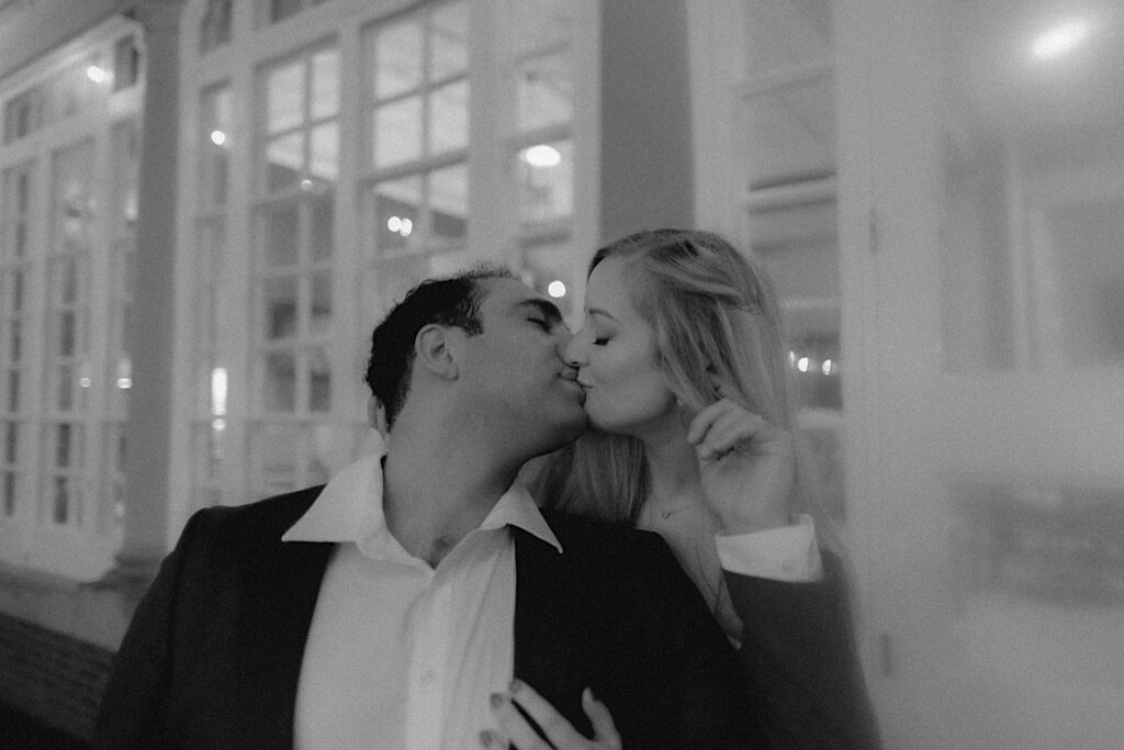 Black and white portrait of woman leaning down behind a man and kissing him from behind in front of lit up windows.