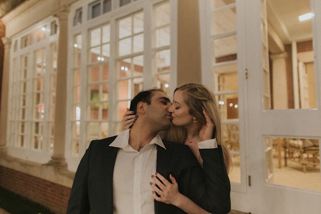 Woman leans down behind a man and kisses him from behind in front of lit up windows.