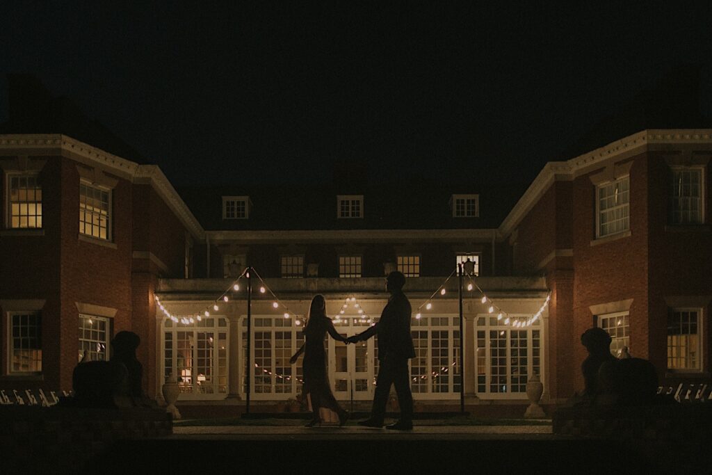 Couple's silhouettes walking along pathway in front of gorgeous mansion building lit up by patio lights at Allerton Park. 