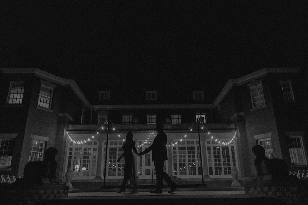 Black and White portrait of couple's silhouettes walking along pathway in front of gorgeous mansion building lit up by patio lights at Allerton Park. 