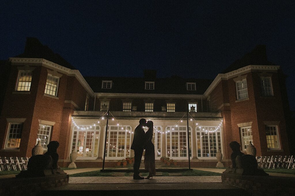 Couple's silhouettes are shown in front of gorgeous mansion building lit up by patio lights at Allerton Park. 