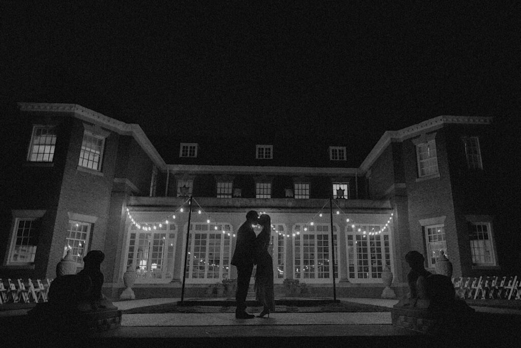 Black and White portrait of couple's silhouettes are shown in front of gorgeous mansion building lit up by patio lights at Allerton Park. 