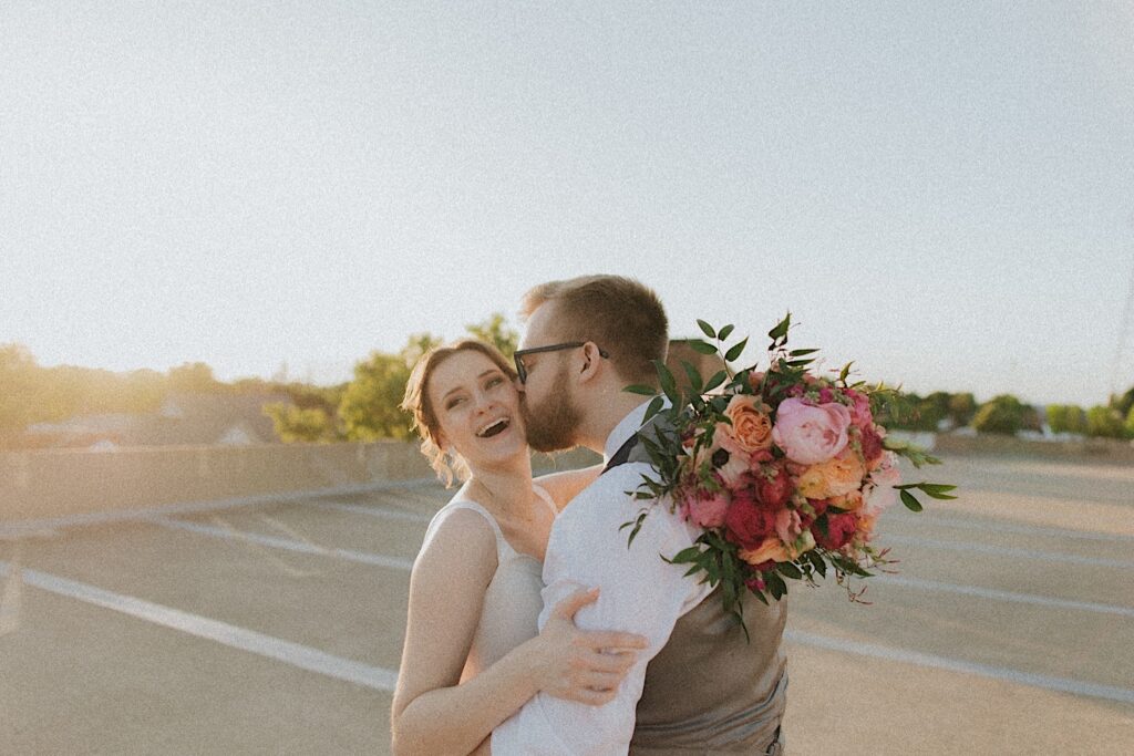 Groom kisses bride as they hug and she puts her bouquet around his neck as they stand on rooftop of Reality on Monroe. 