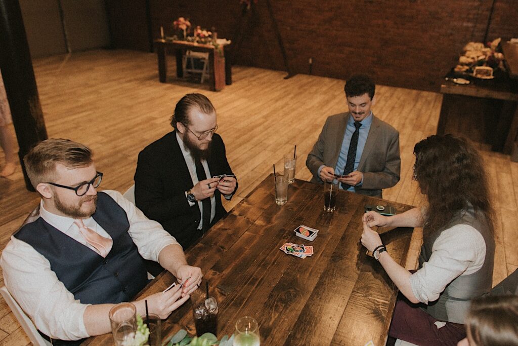 Guests sit around a large wooden table and play uno together with cocktails in front of them during laid back wedding reception in hall at Reality on Monroe. 