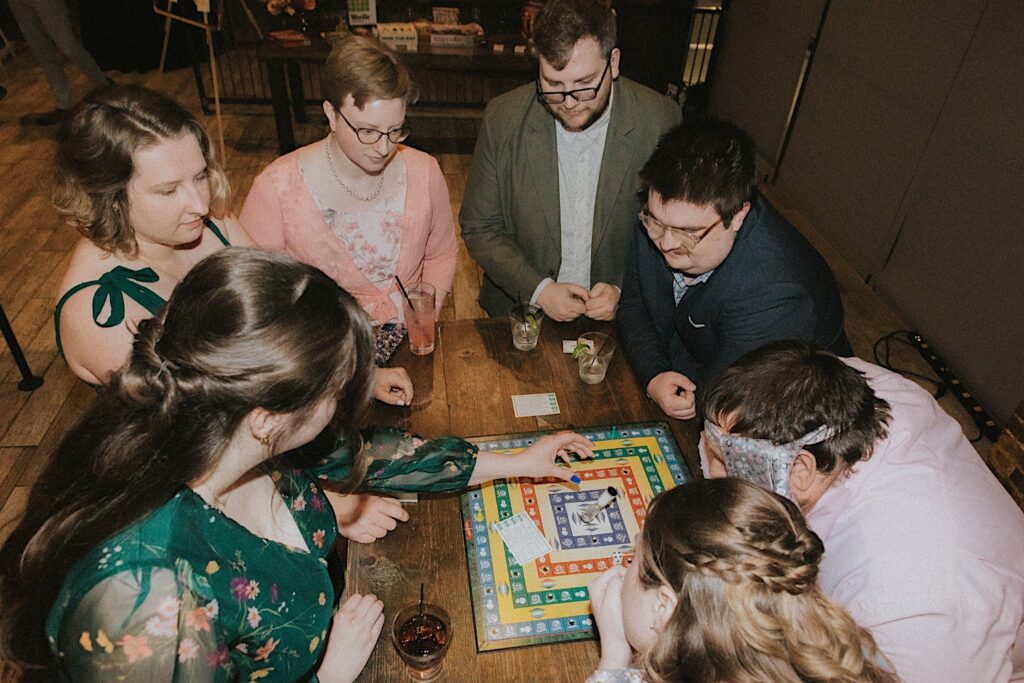 Guests of the bride and groom gather around a table and play board games with drinks in front of them during laid back wedding reception. 