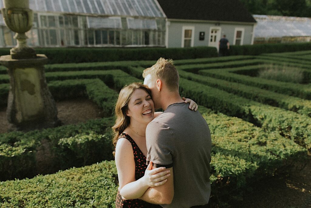 A woman smiles while a man kisses her cheek as they have their engagement photos taken in the gardens of Allerton Park in Champaign, Illinois