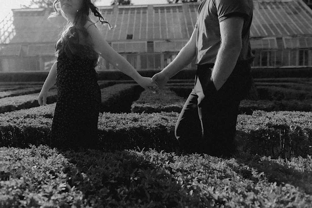 Black and white engagement photo of a couple walking while holding hands through the gardens of Allerton Park in Champaign, Illinois