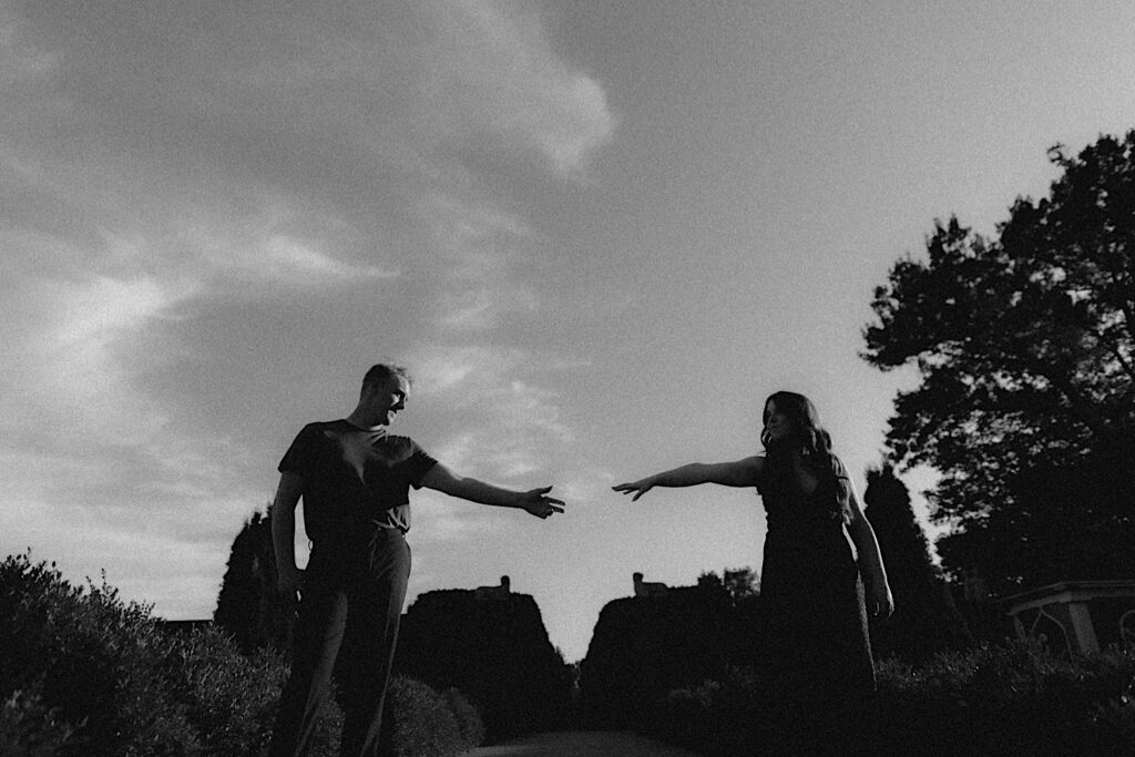 Black and white artful photo of a man and woman reaching their hands towards each other while having their engagement photos taken in Allerton Park of Champaign, Illinois