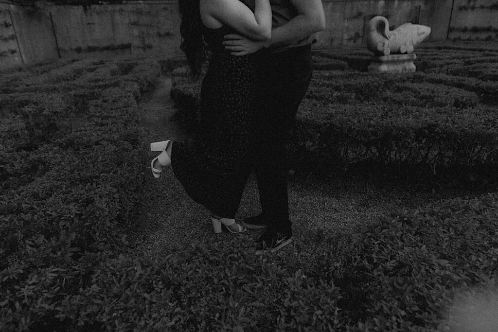 A woman lifts her leg while kissing a man as they stand in a garden of Allerton Park in Champaign, Illinois having their engagement photos taken