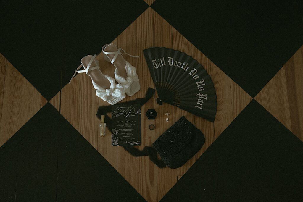 White heels sit on a checkerboard floor next to a fan that says Till Death Do Us Party, black wedding invitations and a black purse for a wedding detail shot. 