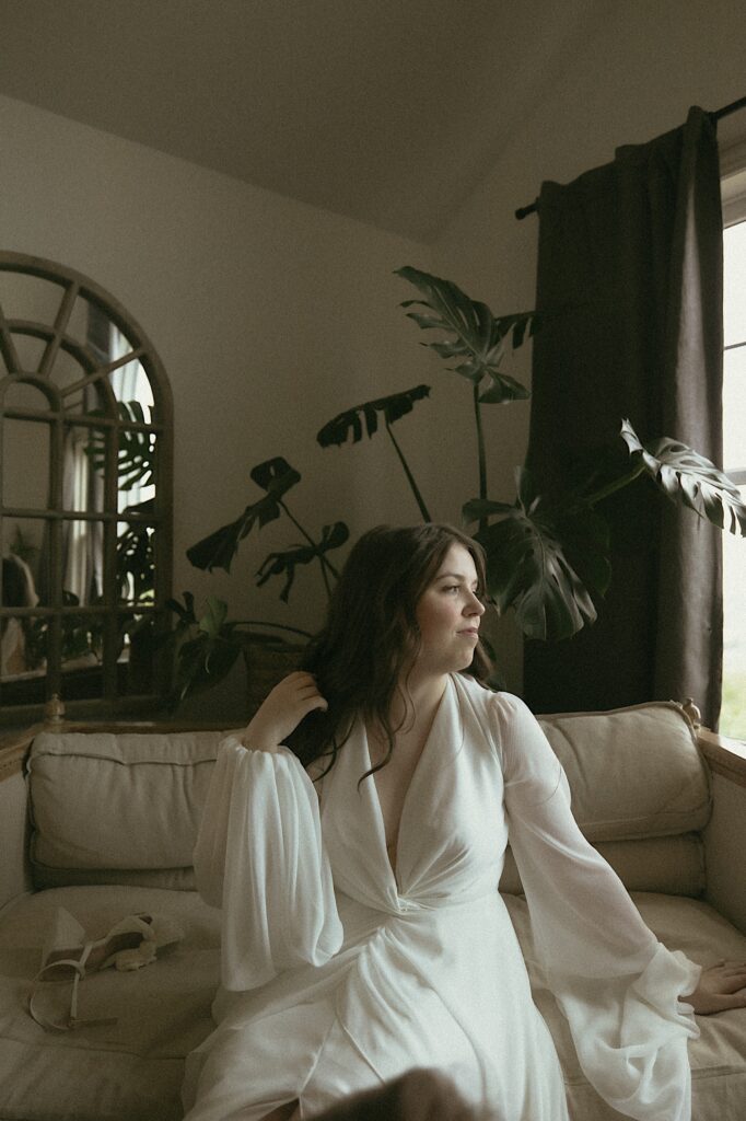 Bride sits on a white couch and flips her hair behind her ear while in a lounge getting ready before wedding ceremony in Washington, Illinois.