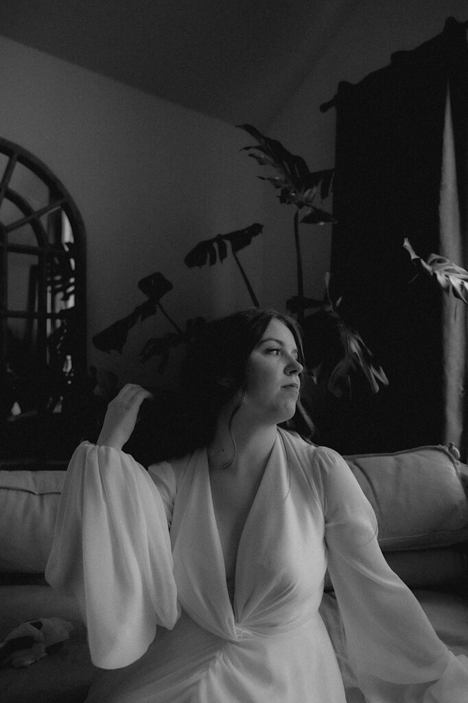 Black and white portrait of Bride sitting on a white couch and flips her hair behind her ear while in a lounge getting ready before wedding ceremony in Washington, Illinois.