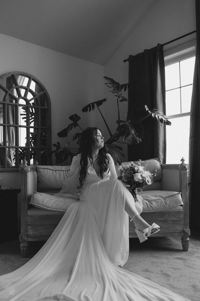 Bride sits with her legs crossed on a white couch and looks outside the window in a beautiful lounge smiling at the camera for a beautiful candid portrait in Illinois.