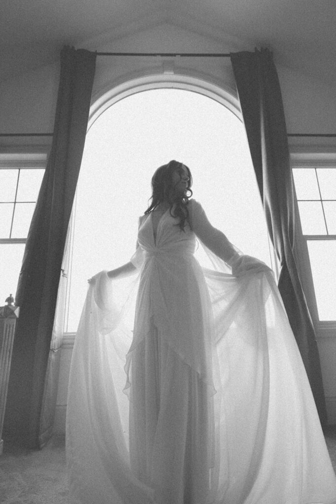 Bride stands in front of a window holding her skirt out for a romantic moody portrait by central Illinois wedding photographer. 