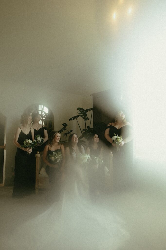 Central Illinois wedding photographer captures moody, film style portrait of bride sitting next to her bridesmaids wearing all black. 