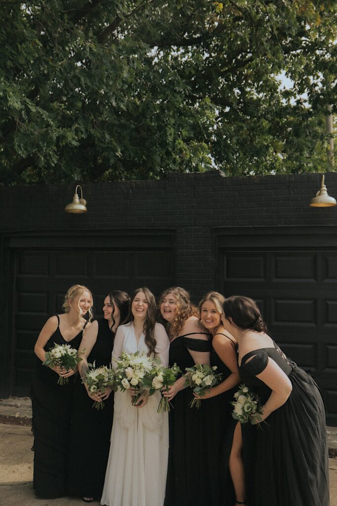 Bride and bridesmaids stand in front of a black garage and candidly laugh.