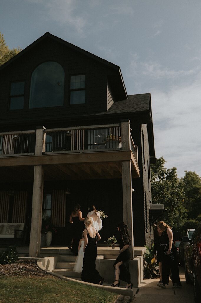 Bridesmaids in all black hold bride's dress as they walk into a lodge style house before wedding ceremony. 