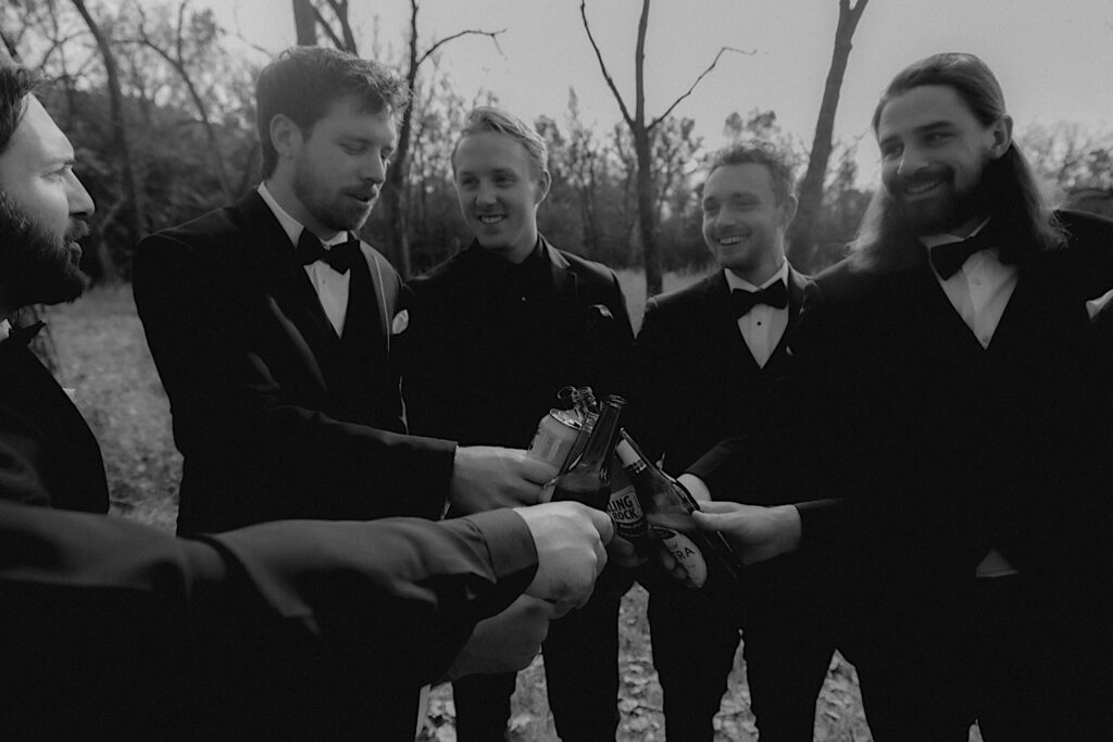 Groomsmen stand in a circle and cheers each other with glass beer bottles in celebration before wedding ceremony. 