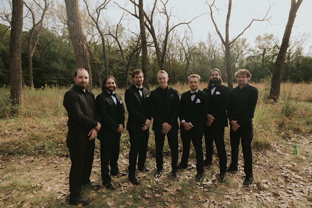 Groomsmen stand around groom wearing all black in a wooded area during autumn. 