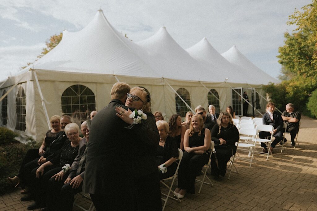 Mother of the groom hugs the groom as he stands at the end of the aisle waiting for the procession during outdoor ceremony in Washington, Illinois. 