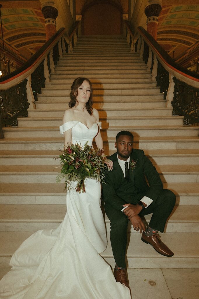 A bride stands in her wedding dress on the marble steps of the Illinois State Capitol Building holding a fall bridal bouquet of mostly greenery next to her husband in a dark green suit. 