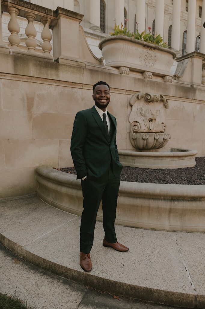 A groom stands outside of the Illinois Capitol building in a dark green suit and black tie smiling at the camera and waiting for his wife to arrive on their wedding day.  