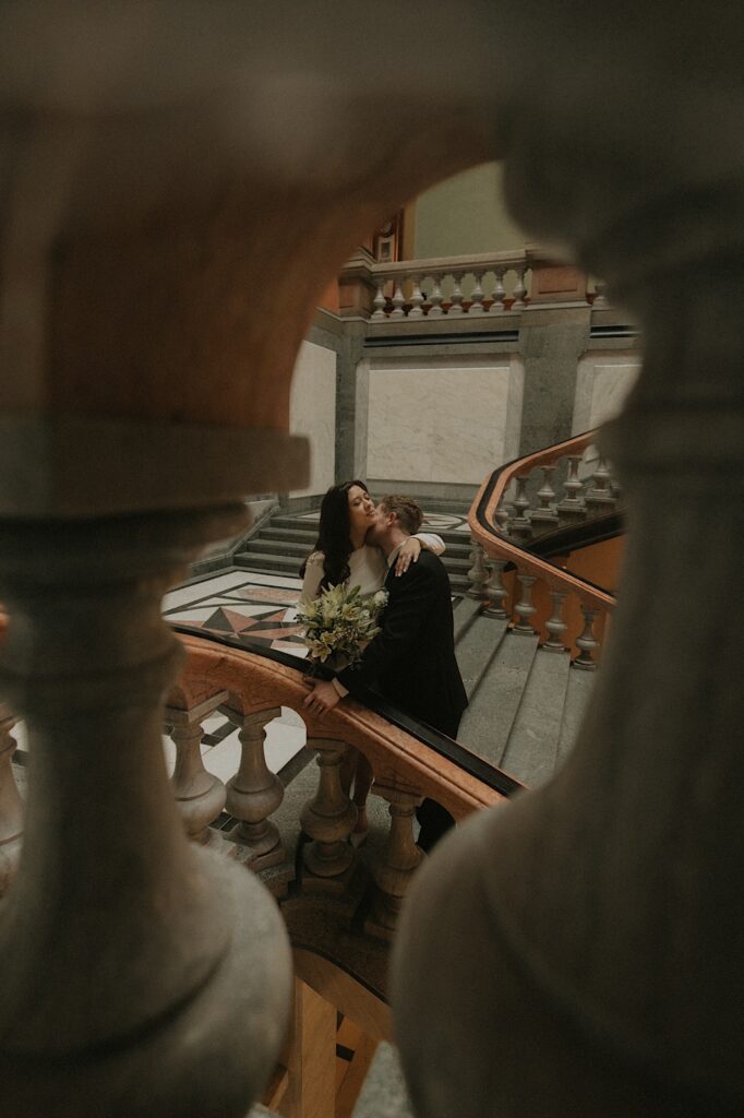 A bride and groom kiss on the marble steps leading up the Illinois State Capitol Building.  The photo is taken from between two marble pillars.