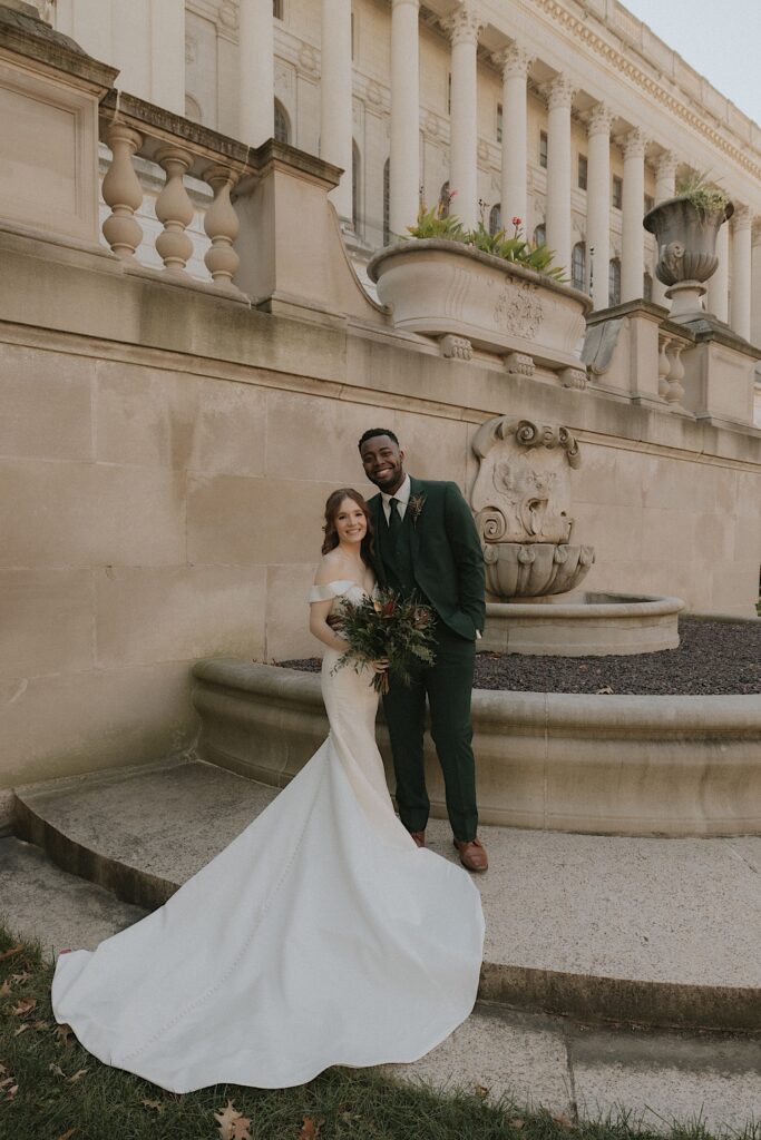 A bride and groom smile for a portrait outside the Illinois State Capitol Building.  The bride is holding a bouquet of greens and wearing a simple elegant white satin dress with a long train.  