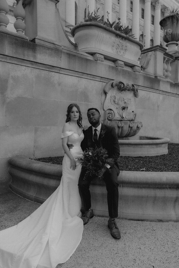 A bride and groom stoically look at the camera for a portrait.  The groom is sitting on the edge of a planter at the Illinois State Capitol building and his bride stands next to him with her hand on his shoulder. 