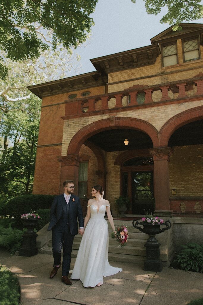 A bride and groom walk down the steps of the Vrooman Mansion in Normal Illinois.  The bride is holding a bouquet of pink flowers. 