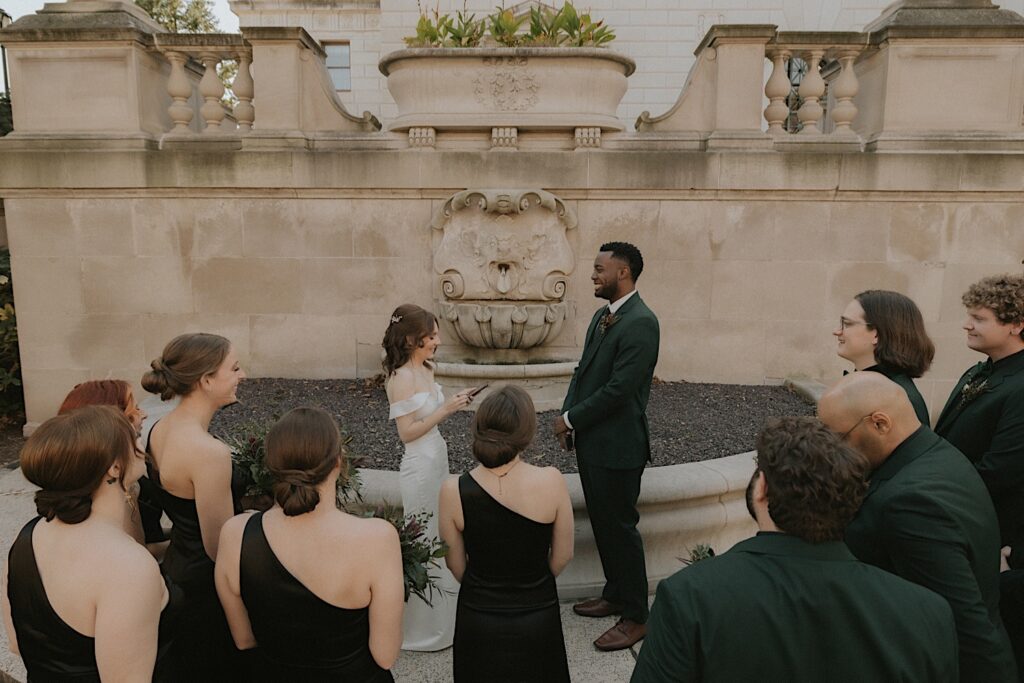 A bride and groom stand in front of a fountain at the Illinois State Capitol building surrounded by their closest friends as they exchange their vows.  Their wedding party wears black dresses and dark green suits. 