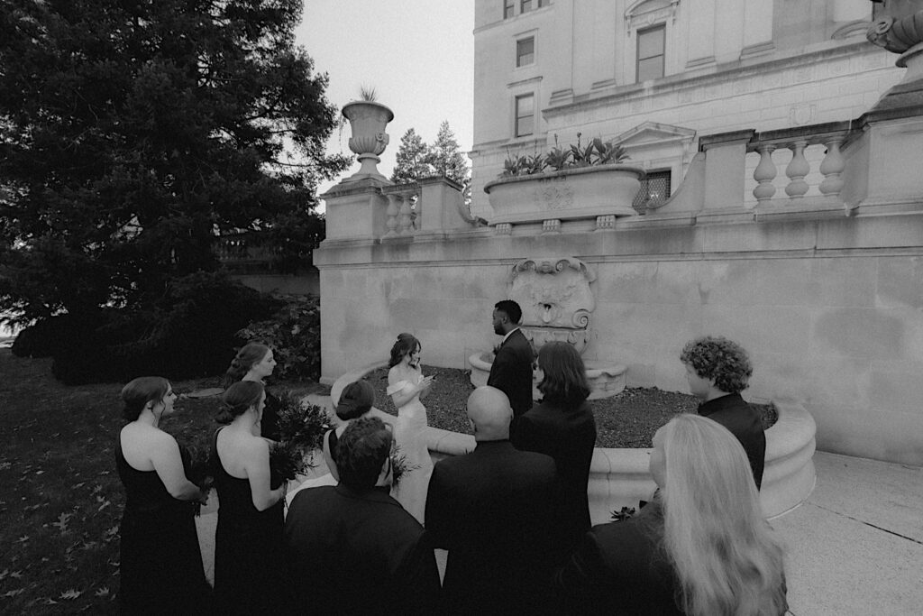 A bride and groom stand in front of the Illinois State Capitol sharing their vows surrounded by their wedding party. 