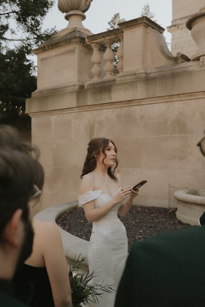 A bride stands in front of her groom reading vows from her phone out in front of the Illinois State Capitol Building.  She is wearing an elegant satin gown and her hair is down in loose curls.  