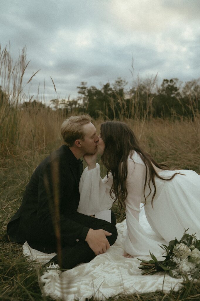 A bride and groom sit on a blanket surrounded by tall grasses on their wedding day.  The bride is wearing a billowing dramatic dress made of organza. 