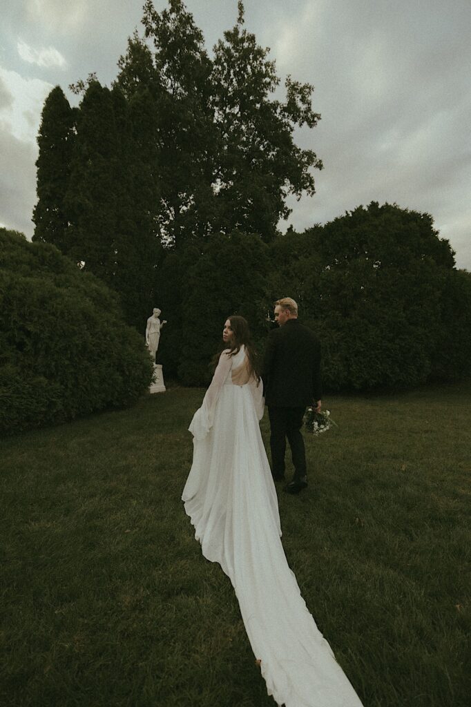 A bride and groom hold hands walking along the grass of Allerton Park in Central Illinois. The brides dress has a train that follows behind them all of the way out of the camera frame and they're walking towards a marble statue on the lawn.