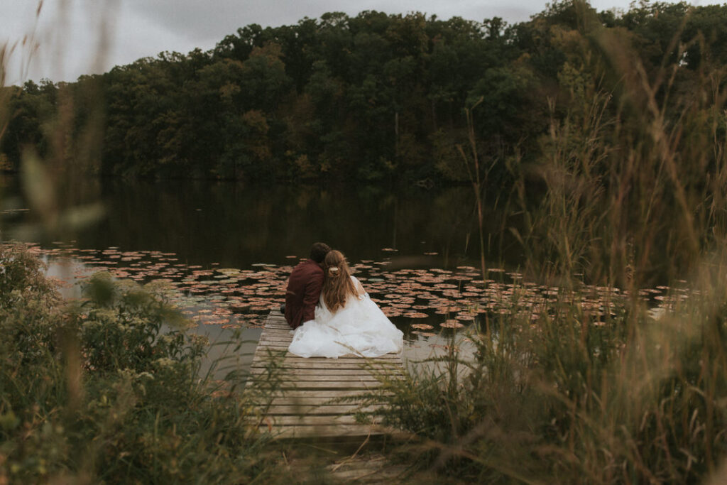 A bride & groom sit on a dock in October after their elopement, surrounded by nature.
