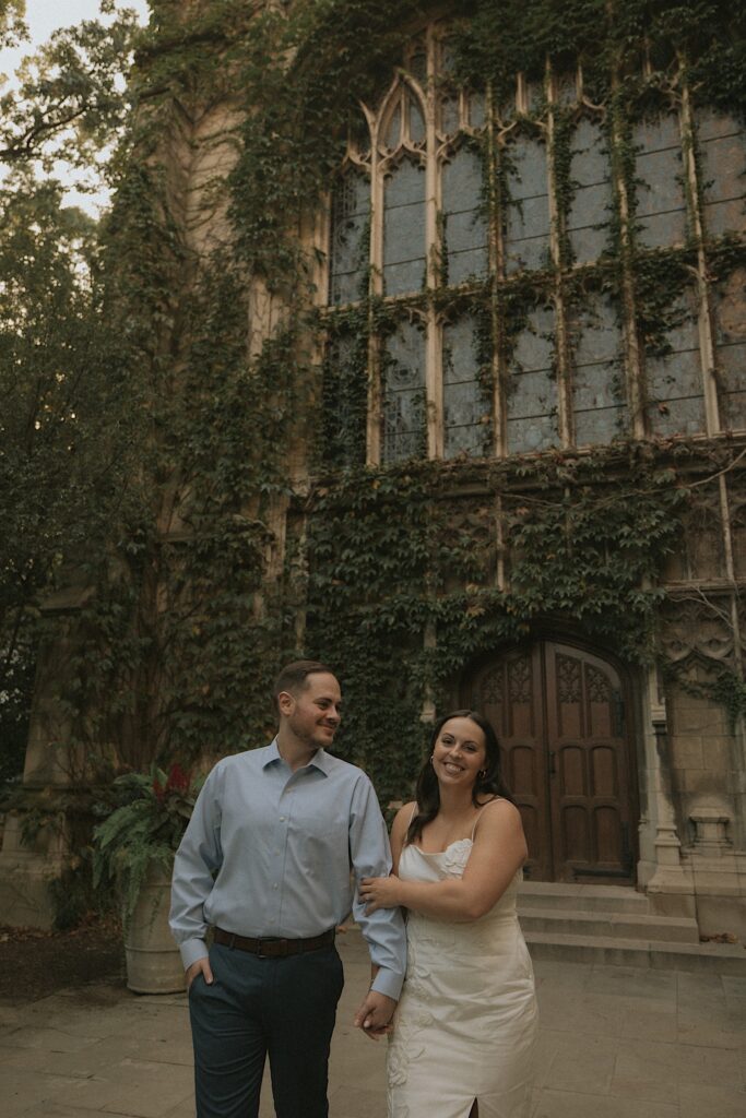 A couple stands holding hands in front of an ornate building on the University of Chicago's Campus for their engagement session