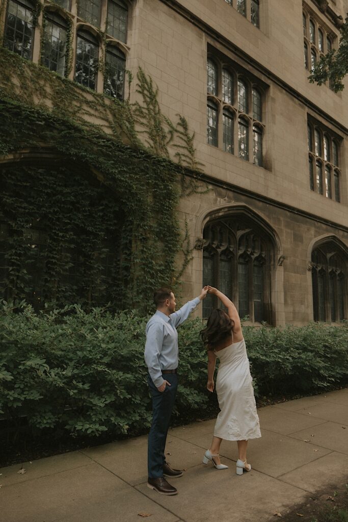 A bride and groom dance outside University of Chicago on a summer day during their engagement session.  The bride wears a cotton dress with white heels and the groom wears blue. 