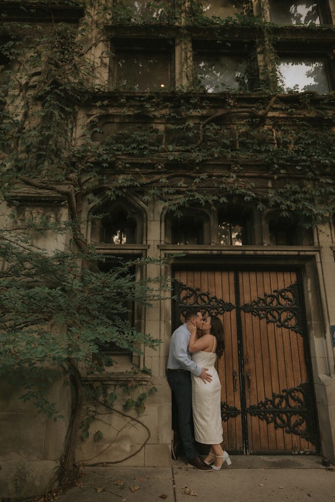 A couple kisses in front of an ornate door of a building covered in Ivy on the University of Chicago's campus. 