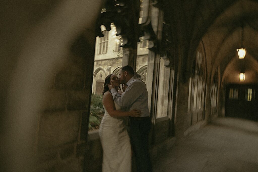 A couple stands under a covered walk way on University of Chicago's campus overlooking a courtyard.   The bride wears a tea length white dress and the groom wears a blue button up shirt. 