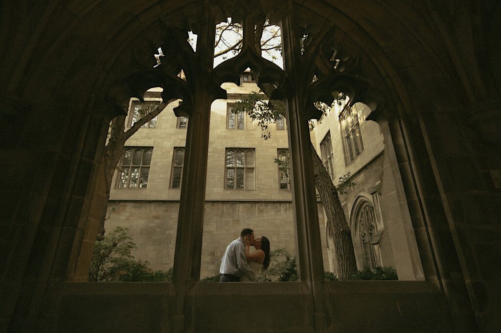 A couple stands under a covered walk way on University of Chicago's campus overlooking a courtyard.   The bride wears a tea length white dress and the groom wears a blue button up shirt. 