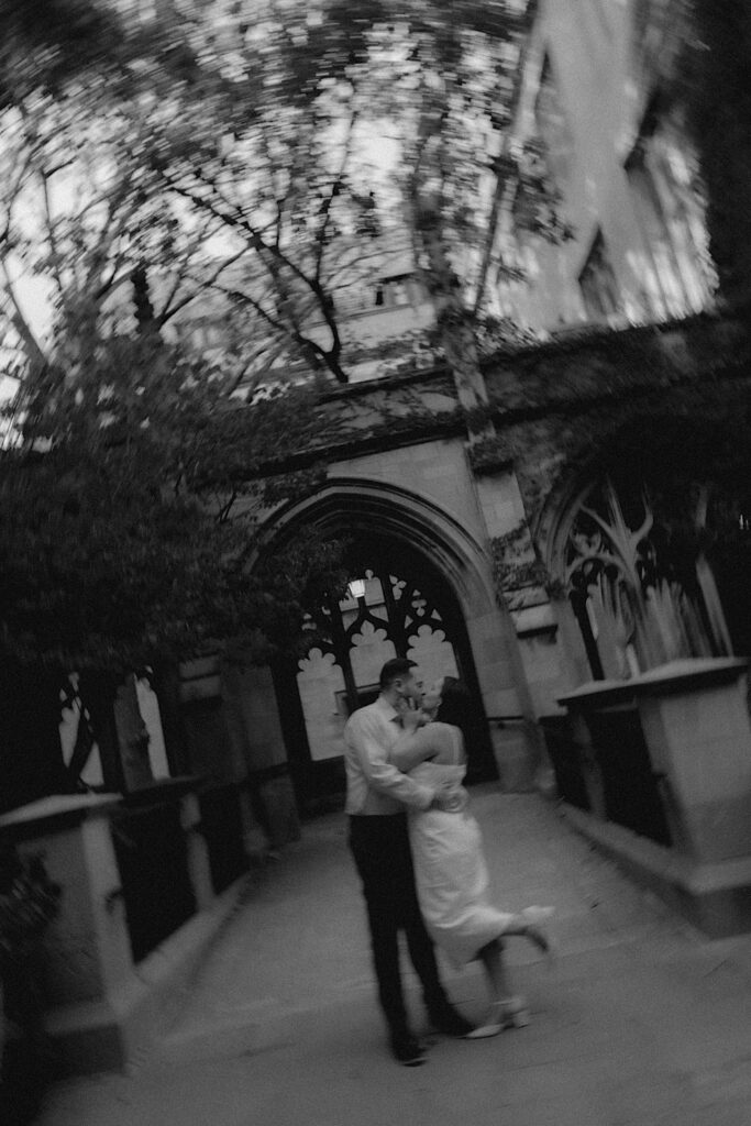 A bride and groom hold one another and kiss outside University of Chicago. You can see ornate windows in the background and the photographer is twisting the camera to make a blurred motion. 