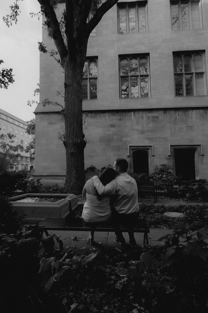 A bride and groom hold one another and kiss outside University of Chicago. They're sitting on a bench in the gardens.