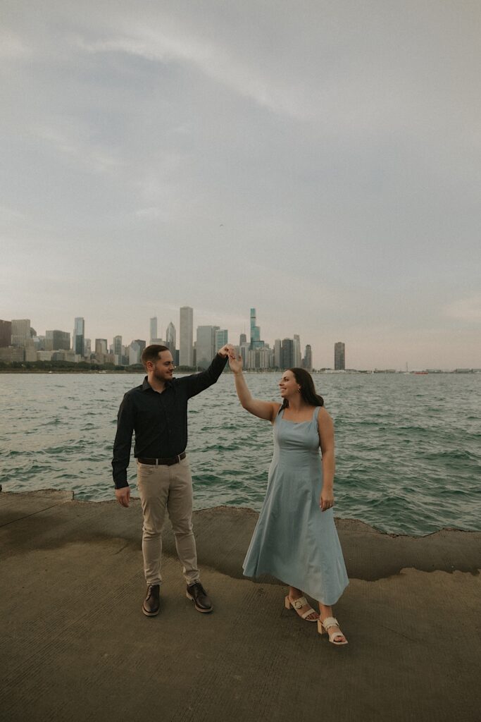 A couple dances overlooking Lake Michigan in Chicago.  The bride wears a blue dress and white slip on heels matching the color of the water with her dress for their engagement session.  